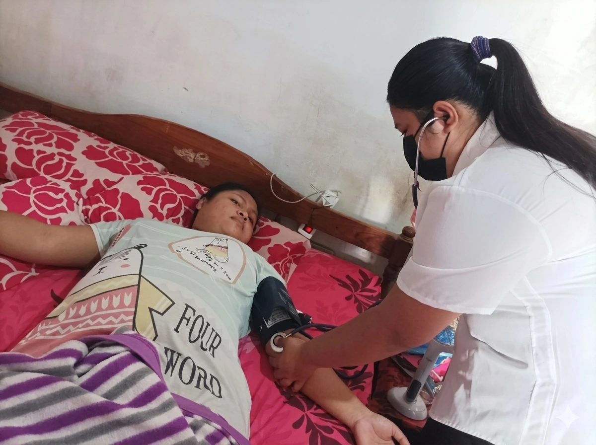 A Bali Sehat nurse in a white uniform using a blood pressure cuff to monitor a pregnant patient lying on a bed at home in Bali, as part of the clinic's free prenatal home visit service.