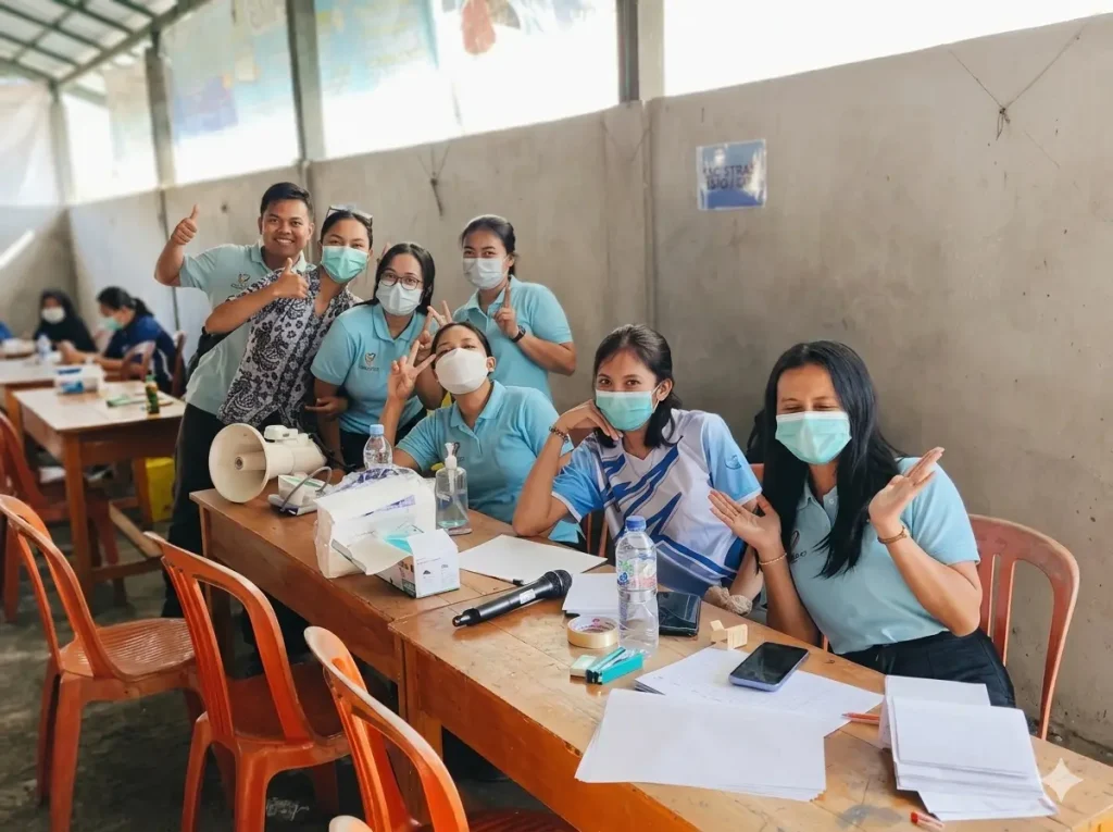 A cheerful group of Bali Sehat medical volunteers and staff in light blue uniforms posing together at their registration desk during the 27th community outreach event in Tiying Tali, Bali.