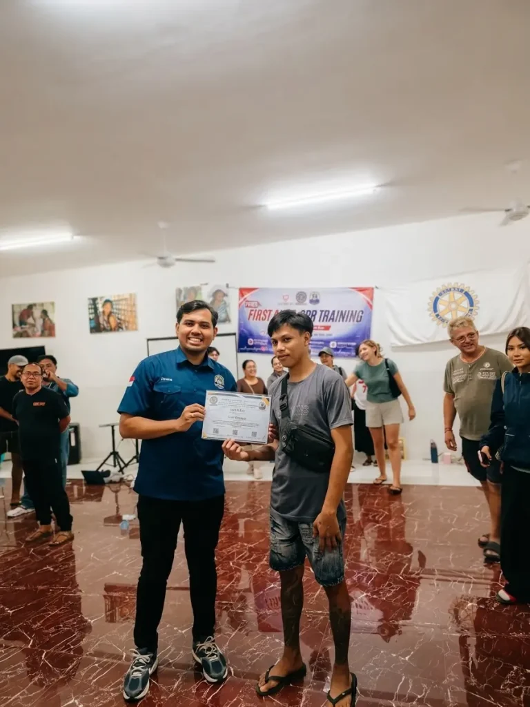 A Bali Sehat team member presents a training certificate to a participant from the Amed community in front of the Rotary International banner.